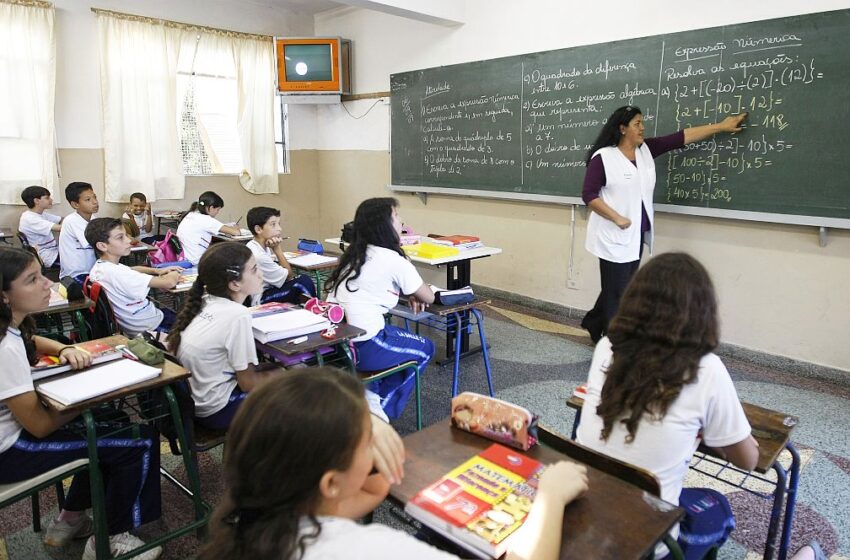 Sala de aula em uma escola brasileira, onde uma professora escreve no quadro negro enquanto alunos uniformizados prestam atenção à explicação.