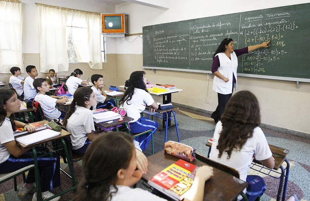 Sala de aula em uma escola brasileira, onde uma professora escreve no quadro negro enquanto alunos uniformizados prestam atenção à explicação.